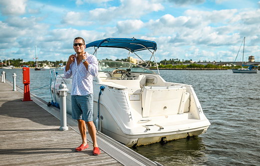 Handsome Latin man in his late 30’s stands on a boat dock in a scenic coastal location. He is next to a sleek looking speed boat. He wears casual cool clothing and has sunglasses and is happy to spend the day boating