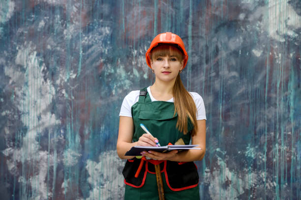 istock Woman in coverall and helmet holding notepad and posing for camera on grey background 1202438093