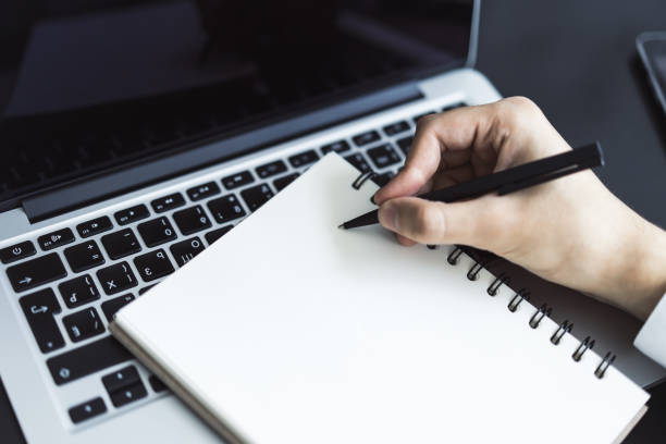 istock Closeup of man hand writing in diary on a table with laptop. Focused on a pen 1202440611