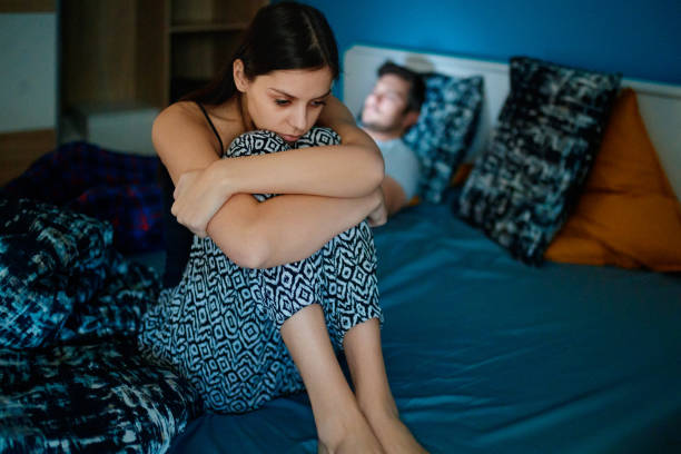 Sad woman sitting on bed with partner in background Sad girlfriend sitting on bed edge, holding her legs after fight, her partner is lying in bed in background, his face is lit by smart phone human relationship stock pictures, royalty-free photos & images