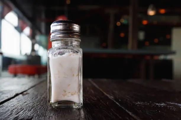 Glass salt shaker on a wooden table with rice grains to stop clogging. Some spilt salt is on the table.