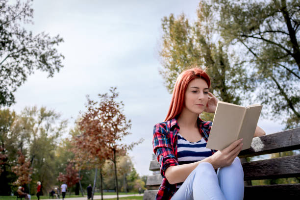 istock Young woman resting on park bench and reading a book. 1202464579