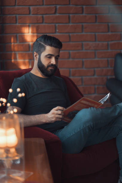 Portrait handsome bearded man wearing casual clothing, sitting in red chair modern loft studio Portrait handsome bearded man wearing casual clothing. Man sitting in red chair modern loft studio, reading comics and relaxing reading comic book stock pictures, royalty-free photos & images