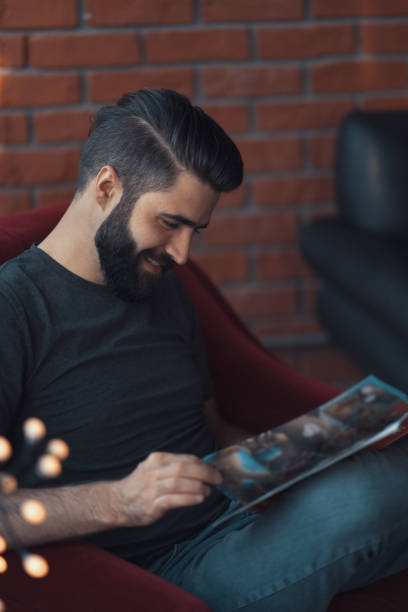 Portrait handsome bearded man wearing casual clothing, sitting in red chair modern loft studio Portrait handsome bearded man wearing casual clothing. Man sitting in red chair modern loft studio, reading comics and relaxing reading comic book stock pictures, royalty-free photos & images