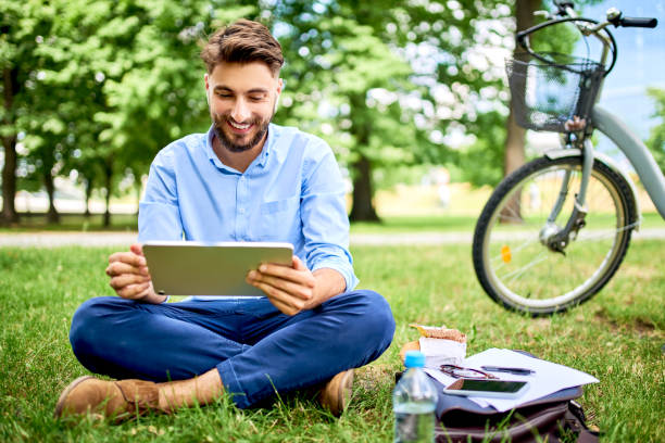 istock Smiling young man sitting in park with tablet and bicycle during lunch break 1202481834
