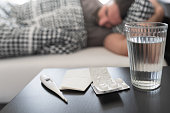 meds, medical thermometer and glass of water on nightstand against person sleeping in bed