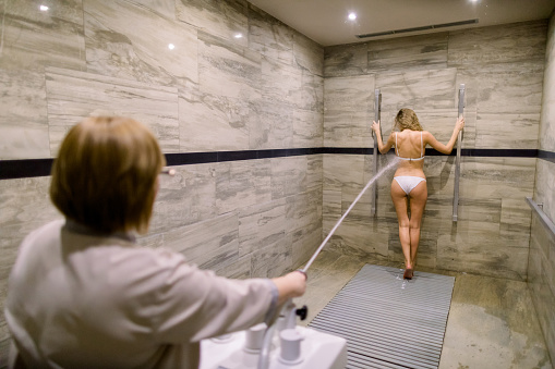 Joven mujer rubia en traje de baño blanco con masaje de alta presión con ducha de Charcot para el tratamiento anticelulítico en el salón de spa photo