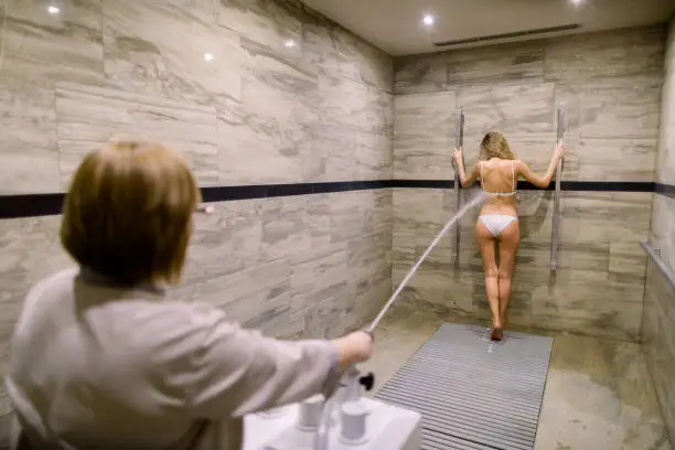 Young blond woman in white swimsuit having high pressure massage with Charcot shower for anti-cellulite treatment in the spa salon.