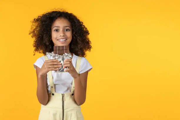 Photo of Little afro girl eating chocolate isolated on yellow background