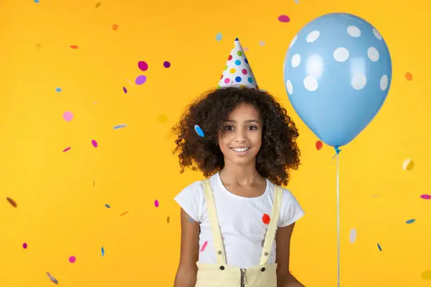 Photo of Funny little lady on birthday party eventwith ballon and confetti falling isolated on yellow background