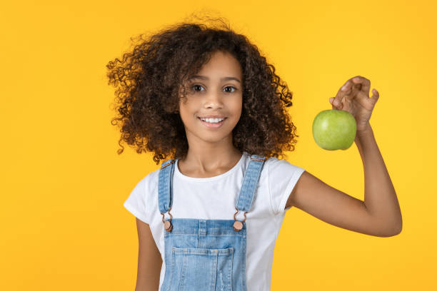 黄色の背景に孤立した緑のリンゴを保持しているかわいいアフリカの女の子。 - child eating apple fruit ストックフォトと画像