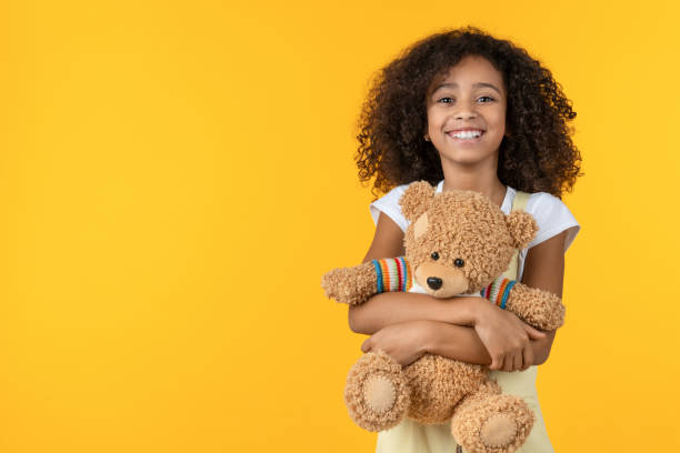 ritratto di ragazza africana sorridente che abbraccia il giocattolo dell'orsacchiotto isolato su sfondo giallo - bambola giocattolo foto e immagini stock