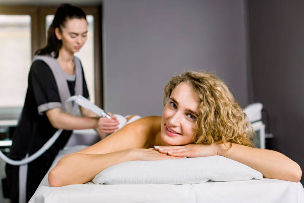 belle femme blonde souriant, regardant l'appareil-photo tout en obtenant le traitement de cavitation d'ultrason par le cosmétologue. procédure de massage anti-cellulite et sous vide au salon de beauté - cellulite beauty treatment health spa lpg photos et images de collection