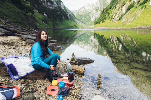 Young woman with long hair in blue jacket resting after her hiking trip near the beautiful mountain lake in Swiss Alps and preparing tasty dinner by gas stove