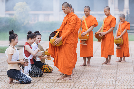 Vang Vieng, Laos - March 2, 2020: A group of monks chant prayers as a woman prays before them after offering alms at dawn, in a local custom called \