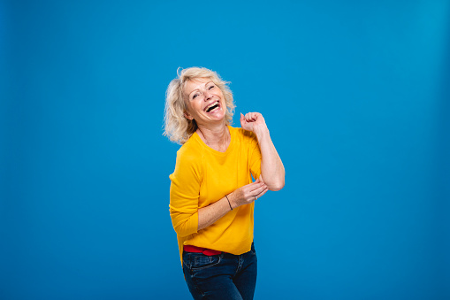 Front view close-up of laughing Caucasian woman in late 50s with medium-length blond hair and wearing long sleeved yellow top and jeans.