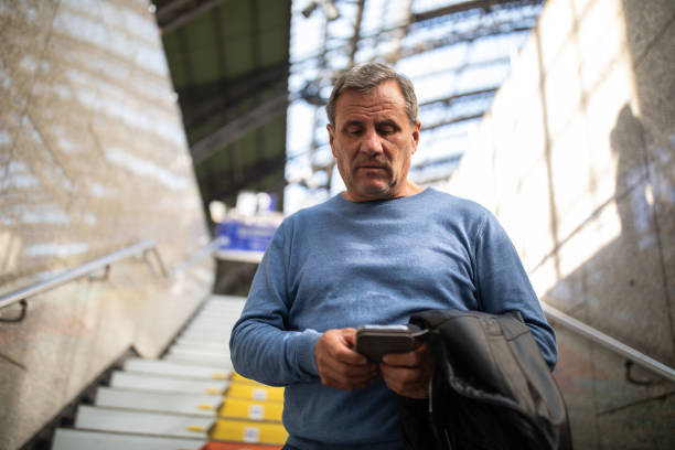 Mature gray-haired male tourist, holding jacket, walking down the stairs at Cologne Central station and using mobile phone