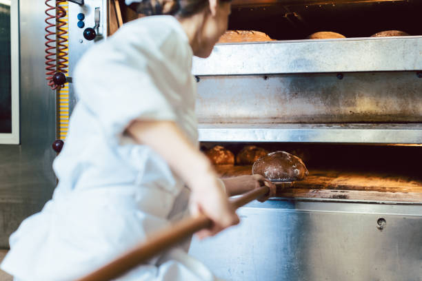 Baker removing two loaves of bread from a professional oven using a wooden peel in a bakery