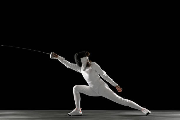 Teen girl in fencing costume with sword in hand isolated on black background Catching moment. Teen girl in fencing costume with sword in hand isolated on black background. Young female model practicing and training in motion, action. Copyspace. Sport, youth, healthy lifestyle. face guard sport stock pictures, royalty-free photos & images