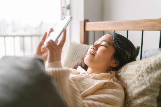 Young and beautiful woman using smart phone in bed in the morning A young and beautiful woman is lying in bed and using a smart phone happily in her bed in the morning. asian checking phone in the morning stock pictures, royalty-free photos & images
