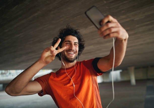 istock Portrait of a smiling young and handsome man with mobile smartphone pulling a peace sign and taking a selfie 1202537124