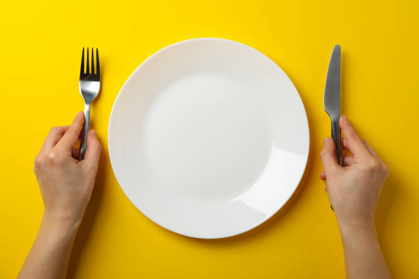 istock Female hands hold fork and knife on yellow background with plate, top view 1202543004