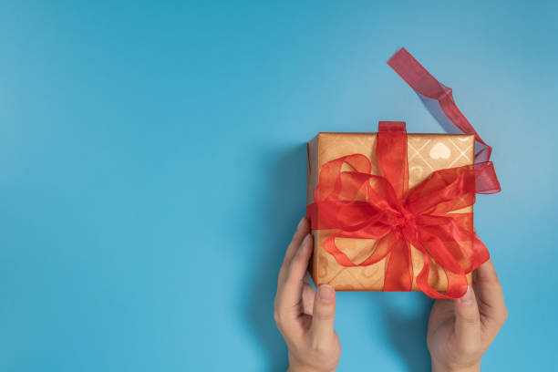 istock Someone hands holding a big gift box tied with red ribbon over blue background 1202547077