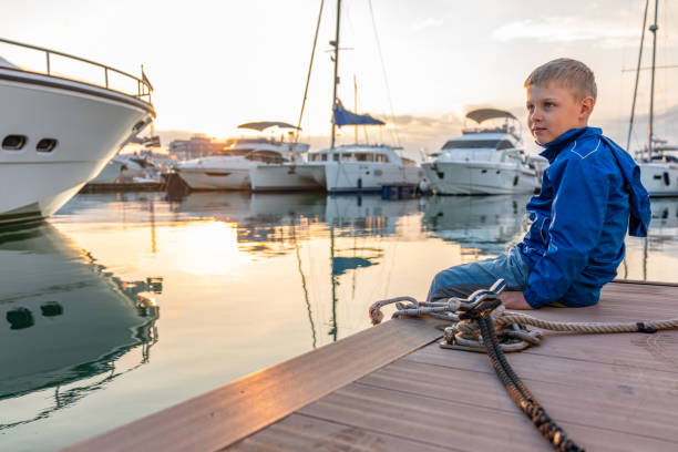 istock A boy in a blue jacket sits on a pier with yachts during sunset. 1202556020