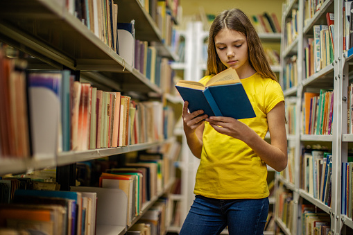 Teenage girl standing in library between bookshelves and reading book. She looks very pensive and cute