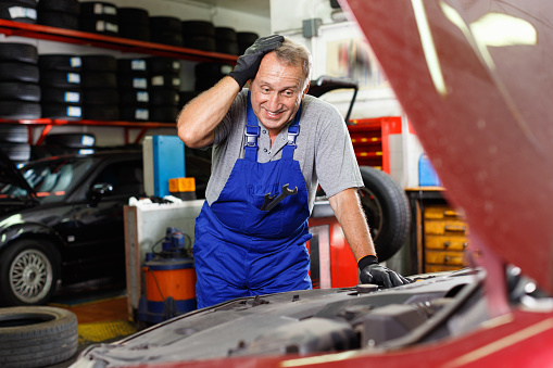 Puzzled  cheerful positive smiling  male mechanic inspecting broken car and determining scope of work at auto service