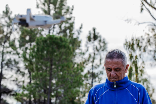 istock Side view of old man flying the drone looking at the remote control while standing against the trees in the park 1202574754