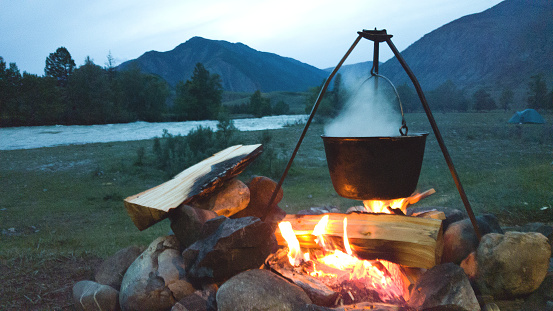 Evening campfire during a camping trip. Cauldron on a burning fire. The tent is far away. Cozy Hiking evening of travelers by the river.