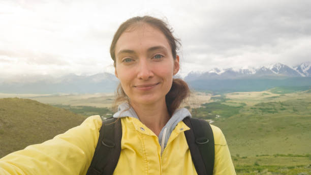 A brunette girl in a bright yellow jacket and with a backpack takes a selfie against the background of the beautiful Altai mountains stock photo