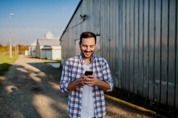 istock Young smiling unshaven caucasian farmer standing next to barn and using smart phone for texting. 1202584764