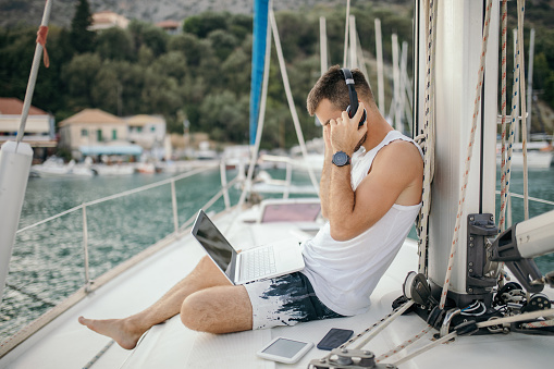 Young man on vacation on yacht using laptop. He sitting in deck of sailboat docked in marina.
