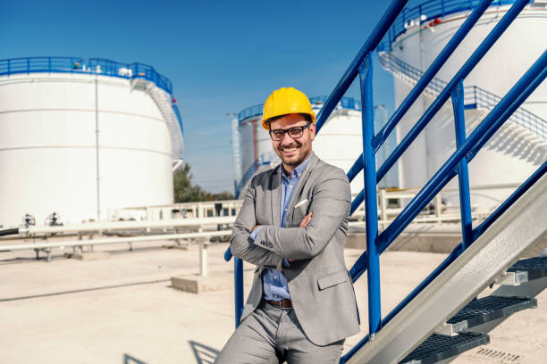 istock Young proud successful refinery owner in suit with helmet on head leaning on railing and holding arms crossed while looking at camera. In background are oil tanks. 1202585642