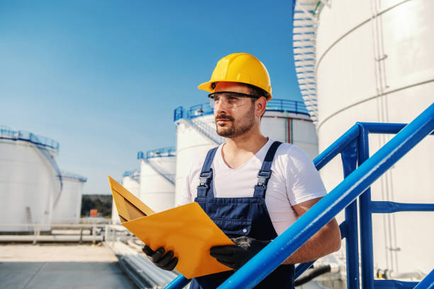 istock Young hardworking Caucasian unshaven refinery worker in working clothes with helmet on head and protective glasses holding folder with documents. 1202586332