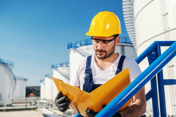 istock Young hardworking Caucasian unshaven refinery worker in working clothes with helmet on head and protective glasses holding folder with documents. 1202586376