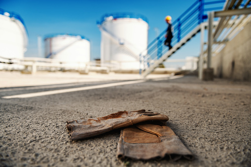 Close up of gloves on ground. In blurry background are businessman who is going down the stairs and tanks with oil. Refinery exterior.