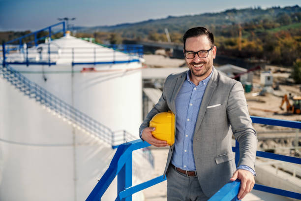 istock Cheerful handsome caucasian smiling refinery owner standing on height, holding helmet under armpit and looking at camera. In background is tank with oil. Refinery exterior. 1202590936