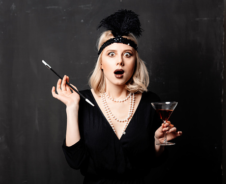 Beautiful blonde woman in twenties years clothes with smoking pipe and cocktail on dark background