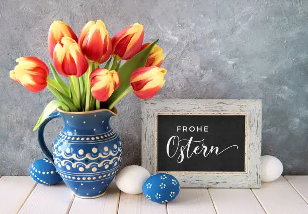 Red-yellow tulips in classic blue ceramic pitcher with Easter eggs and a blackboard on gray background. "Frоhe Ostern" in German means Happy Easter.