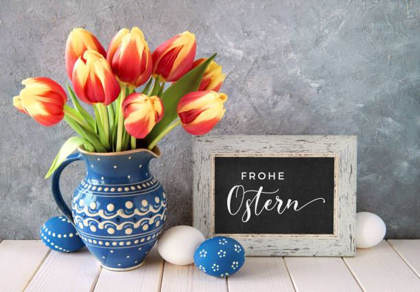 德語中的"fr_he ostern"意為復活節快樂。紅黃色鬱金香在經典的藍色陶瓷罐與復活節彩蛋和黑板，文本 - ostern 個照片及圖片檔