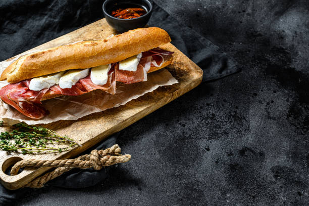 istock Baguette sandwich with prosciutto ham, Camembert cheese on a cutting Board. Black background, top view, space for text 1202596003
