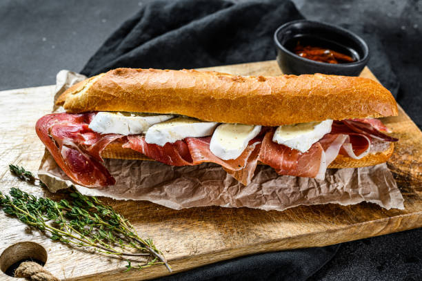 istock Baguette sandwich with prosciutto ham, Camembert cheese on a cutting Board. Black background, top view. 1202596063