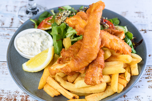 A plate of 'Fish and Chips' with battered fish, potato chips, salad, tartar sauce and lemon wedge.