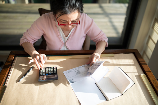 Asian senior woman sitting at desk alone and working with calculator and papers