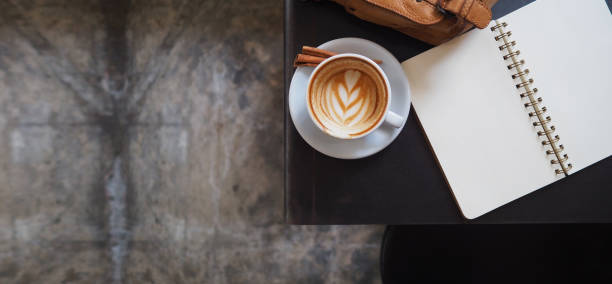 A cup of hot cappuccino coffee with beautiful latte art and open page book and vintage leather bag on table in coffee shop, selective focus for background
