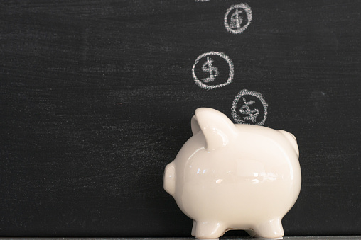 A concept photo of finances and savings.  A ceramic piggy bank sits in front of a black chalkboard with coins hand drawn in chalk coming out of it and/or going into it.  The background is a dark black chalkboard.