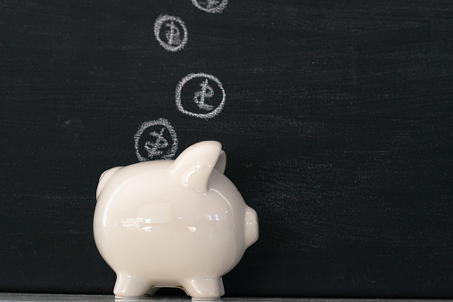 A concept photo of finances and savings.  A ceramic piggy bank sits in front of a black chalkboard with coins hand drawn in chalk coming out of it and/or going into it.  The background is a dark black chalkboard.